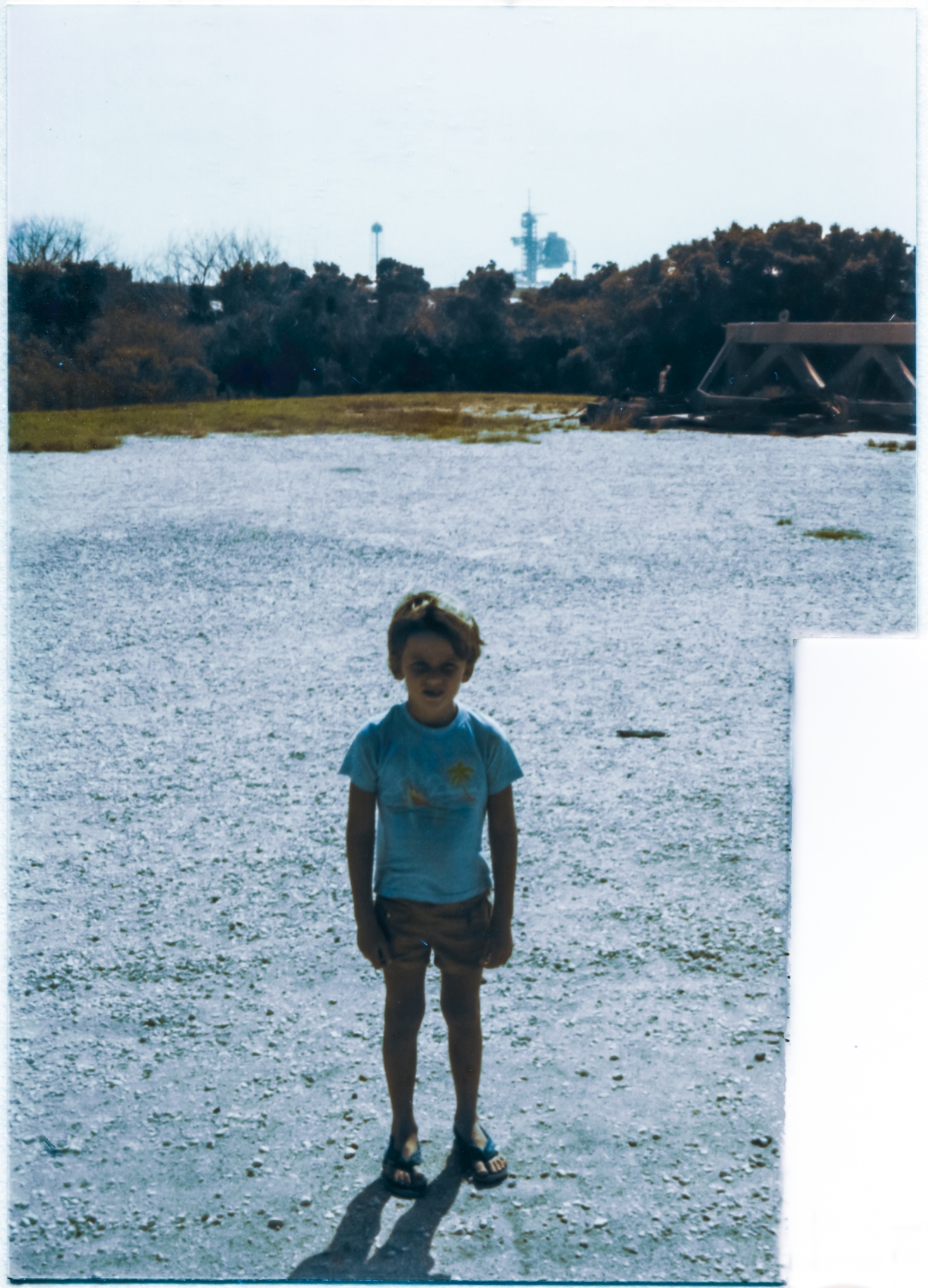 Image 083. Following the landing of STS-41G and the departure of the crowd there viewing it, a six-year-old Kai MacLaren stands backlit on the bleak gravel expanse of the empty parking lot just south of the Ops Building at Space Shuttle Launch Complex 39-B, Kennedy Space Center, Florida. In the far distance behind him, you can see the SSW Water Tower, FSS, and RSS of Pad A standing above the horizon, where Mission 41-G had departed from only eight days previously. Photo by James MacLaren.